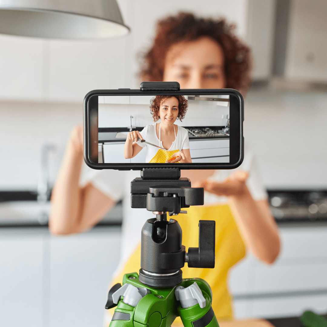 Woman cooking in her kitchen, filming her episode on a mobile phone on a tripod.